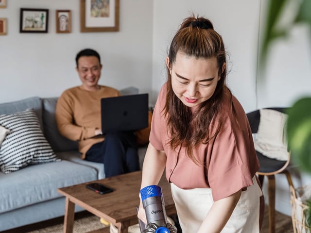 Woman holding a handheld canister vacuum attachment in a living room with a man using a laptop on the couch behind her.