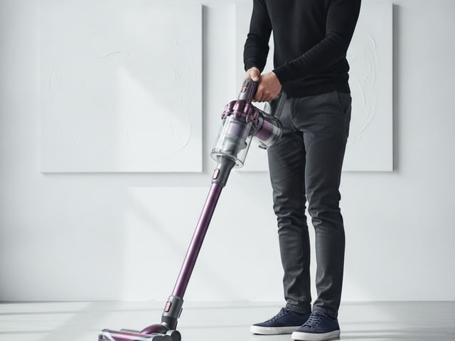 Cordless stick vacuum with transparent collection tank being held upright by person wearing dark clothing in minimalist room setting.