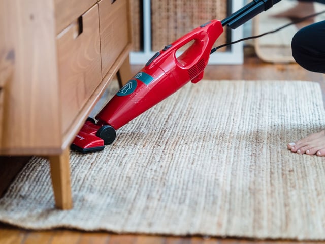 Red handheld vacuum being used to clean under wooden furniture on a light-colored area rug.