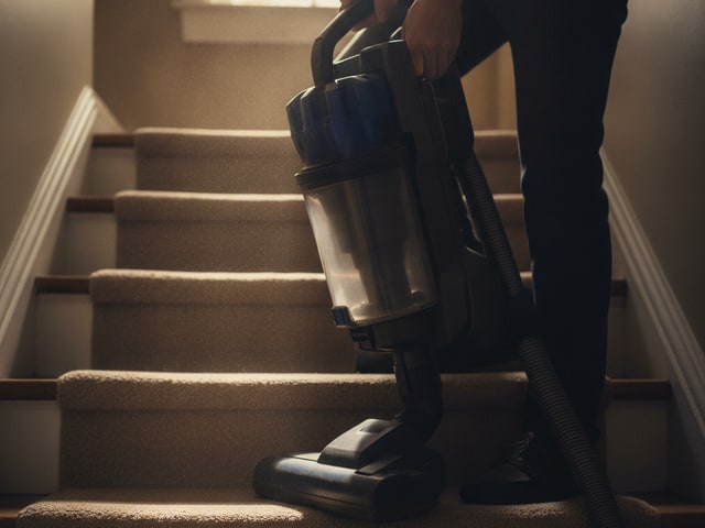 Person's hands straining to lift heavy upright vacuum cleaner up narrow staircase